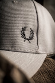 A close-up of a textured fabric cap featuring an embroidered laurel wreath design. The lighting creates a soft shadow effect, highlighting the intricate stitching and the cap's material.