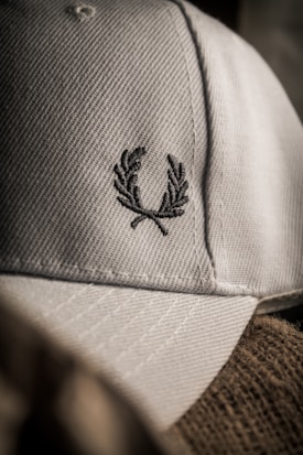 A close-up of a textured fabric cap featuring an embroidered laurel wreath design. The lighting creates a soft shadow effect, highlighting the intricate stitching and the cap's material.