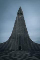 A tall, imposing concrete church with a distinctively modern, geometric design and a clock near the top. The building's facade features a series of vertical columns that taper as they reach the pointed spire. The sky is overcast, contributing to a somber atmosphere.