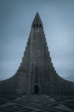 A tall, imposing concrete church with a distinctively modern, geometric design and a clock near the top. The building's facade features a series of vertical columns that taper as they reach the pointed spire. The sky is overcast, contributing to a somber atmosphere.