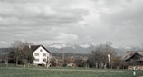 A rural landscape features a house with a traditional design and a red-tiled roof, set against a backdrop of snow-capped mountains. A Swiss flag is visible near the house, surrounded by bare trees and a green field. The sky is overcast with thick, textured clouds.