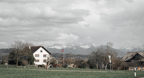 A rural landscape features a house with a traditional design and a red-tiled roof, set against a backdrop of snow-capped mountains. A Swiss flag is visible near the house, surrounded by bare trees and a green field. The sky is overcast with thick, textured clouds.
