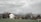 A rural landscape features a house with a traditional design and a red-tiled roof, set against a backdrop of snow-capped mountains. A Swiss flag is visible near the house, surrounded by bare trees and a green field. The sky is overcast with thick, textured clouds.