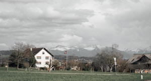 A rural landscape features a house with a traditional design and a red-tiled roof, set against a backdrop of snow-capped mountains. A Swiss flag is visible near the house, surrounded by bare trees and a green field. The sky is overcast with thick, textured clouds.