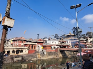 A historical and cultural site featuring traditional architecture with tiered rooftops. The scene includes several buildings along a riverside, some smoke or mist near the buildings, and a bright sky. People are present along the riverbank, with some utility poles and wires visible.