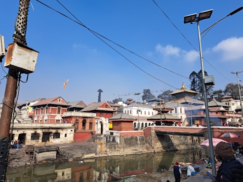 A historical and cultural site featuring traditional architecture with tiered rooftops. The scene includes several buildings along a riverside, some smoke or mist near the buildings, and a bright sky. People are present along the riverbank, with some utility poles and wires visible.