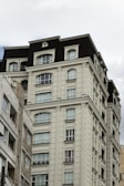 A multi-story residential building with classical architectural elements. The facade features symmetrical windows and ornamental details, painted in a light cream color with contrasting dark roofing. The adjacent building is slightly visible and shares a similar architectural style.
