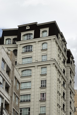 A multi-story residential building with classical architectural elements. The facade features symmetrical windows and ornamental details, painted in a light cream color with contrasting dark roofing. The adjacent building is slightly visible and shares a similar architectural style.
