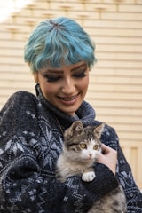 A person with blue hair wearing a dark patterned sweater is smiling while holding a fluffy tabby cat. The background consists of horizontal wooden slats, adding a neutral contrast to the vibrant colors of the person and the cat.