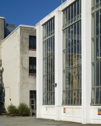 A modern industrial building with large glass windows dominates the scene. The structure is composed of concrete and metal, with visible signs of weathering and age. The windows reflect the clear blue sky, and the building appears to house some type of industrial machinery.