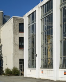 A modern industrial building with large glass windows dominates the scene. The structure is composed of concrete and metal, with visible signs of weathering and age. The windows reflect the clear blue sky, and the building appears to house some type of industrial machinery.