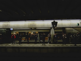 People are standing on a train platform, waiting for a train. The station is dimly lit, and there is a mix of individuals some looking at their phones while others are engaged in conversations. The platform has a modern, industrial look with overhead lighting and signage. A train is visible on the opposite side of the platform.