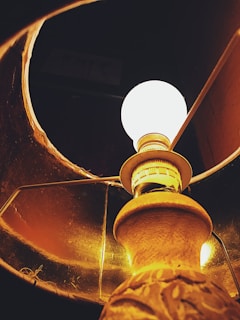 An upward view of a lit lamp with a glowing bulb. The lamp has a decorative base and is encased in a fabric lampshade that casts a warm, amber light. The interior of the lampshade is visible, highlighting its texture and color.