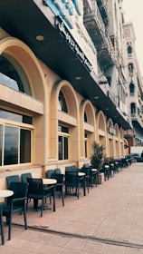 A street with a series of arched windows and outdoor seating in the form of black chairs and round tables lined along the sidewalk. The building features a beige facade with a dark sign in Arabic above. There are potted plants spaced throughout the seating area, and the architecture is reminiscent of classic urban designs.