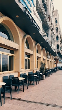 A street with a series of arched windows and outdoor seating in the form of black chairs and round tables lined along the sidewalk. The building features a beige facade with a dark sign in Arabic above. There are potted plants spaced throughout the seating area, and the architecture is reminiscent of classic urban designs.
