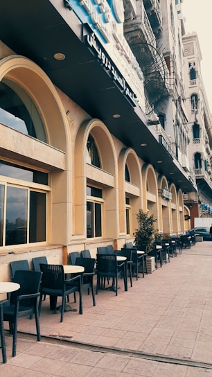 A street with a series of arched windows and outdoor seating in the form of black chairs and round tables lined along the sidewalk. The building features a beige facade with a dark sign in Arabic above. There are potted plants spaced throughout the seating area, and the architecture is reminiscent of classic urban designs.
