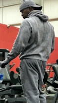 A person is standing in a gym environment, wearing a gray hoodie and sweatpants with a cap worn backwards. Various gym equipment, including a weight bench and weights, are visible in the background.
