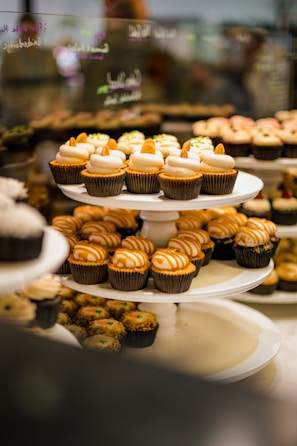 A display of assorted cupcakes arranged on a tiered stand, featuring a variety of designs with icing and toppings. The cupcakes are showcased in a bakery setting with soft, warm lighting, and a reflective glass surface in the background showing faint handwritten words.