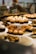 A display of assorted cupcakes arranged on a tiered stand, featuring a variety of designs with icing and toppings. The cupcakes are showcased in a bakery setting with soft, warm lighting, and a reflective glass surface in the background showing faint handwritten words.