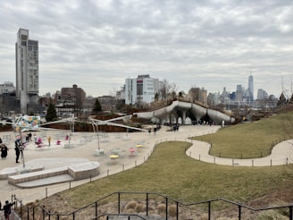 An urban park with modern architectural features and a spacious lawn area. Several people are walking and gathering around paths and open spaces with colorful outdoor seating. The skyline of a city with high-rise buildings is visible in the background under a cloudy sky.