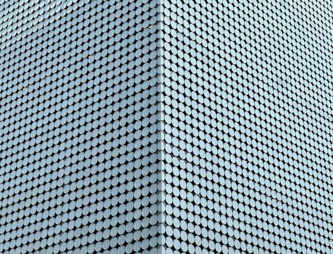 A large facade is covered with a uniform pattern of round, light blue tiles that create a textured, geometric appearance. The tiles are densely packed, aligning in diagonal rows that emphasize the modern architectural design.