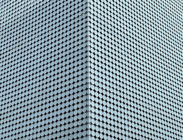A large facade is covered with a uniform pattern of round, light blue tiles that create a textured, geometric appearance. The tiles are densely packed, aligning in diagonal rows that emphasize the modern architectural design.