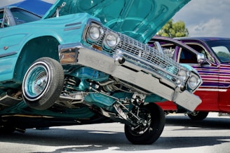 A turquoise lowrider car with its front lifted off the ground, showcasing its hydraulic system. The car's chrome details glisten in the sunlight. Another car, with a dark red and purple striped paint job, is parked nearby. The setting appears to be an outdoor car show or event.