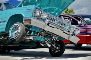 A turquoise lowrider car with its front lifted off the ground, showcasing its hydraulic system. The car's chrome details glisten in the sunlight. Another car, with a dark red and purple striped paint job, is parked nearby. The setting appears to be an outdoor car show or event.