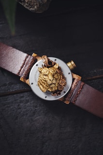 A luxury wristwatch with a leather strap, featuring a detailed golden lion head with a rose on its face within the central dial. The watch is placed on a dark textured surface.