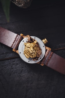 A luxury wristwatch with a leather strap, featuring a detailed golden lion head with a rose on its face within the central dial. The watch is placed on a dark textured surface.