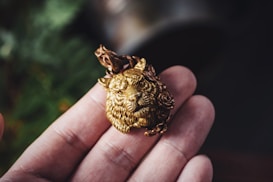 A detailed, golden wolf face pendant held between fingers, with intricate carvings including floral designs around the edges. The background is slightly blurred with hints of greenery.