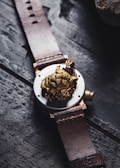 A leather-strapped watch featuring a detailed, golden lion motif on the back of the case. The design includes intricate rose engravings surrounding the lion's head, creating a luxurious and artistic look. The watch is placed on a dark, textured wooden surface.