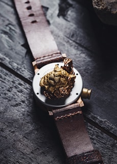 A leather-strapped watch featuring a detailed, golden lion motif on the back of the case. The design includes intricate rose engravings surrounding the lion's head, creating a luxurious and artistic look. The watch is placed on a dark, textured wooden surface.