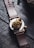 A leather-strapped watch featuring a detailed, golden lion motif on the back of the case. The design includes intricate rose engravings surrounding the lion's head, creating a luxurious and artistic look. The watch is placed on a dark, textured wooden surface.