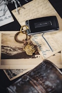 A Jaguar car key fob with a golden lion head keychain sits on top of vintage postcards with cursive writing and a black-and-white photograph of a landscape. The background includes a metallic object and part of a skeleton model's hand.