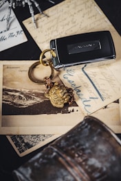 A Jaguar car key fob with a golden lion head keychain sits on top of vintage postcards with cursive writing and a black-and-white photograph of a landscape. The background includes a metallic object and part of a skeleton model's hand.