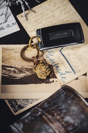 A Jaguar car key fob with a golden lion head keychain sits on top of vintage postcards with cursive writing and a black-and-white photograph of a landscape. The background includes a metallic object and part of a skeleton model's hand.