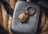 A metallic flask is placed against a backdrop of coiled rope. Resting on the flask is a detailed, gold-colored keychain featuring a lion's head and a circular ring with engraved text. The scene combines rugged textures with sophisticated details, creating a visually intriguing contrast.
