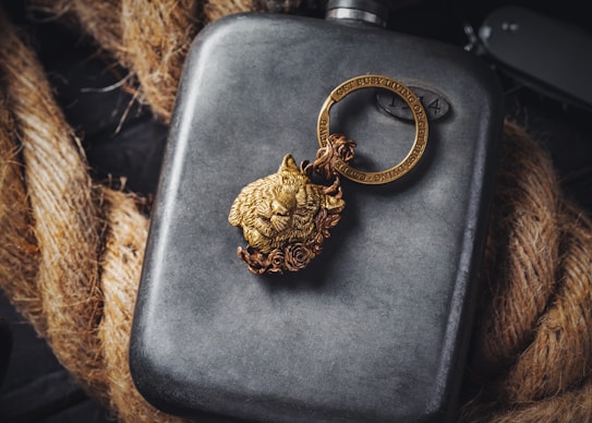 A metallic flask is placed against a backdrop of coiled rope. Resting on the flask is a detailed, gold-colored keychain featuring a lion's head and a circular ring with engraved text. The scene combines rugged textures with sophisticated details, creating a visually intriguing contrast.