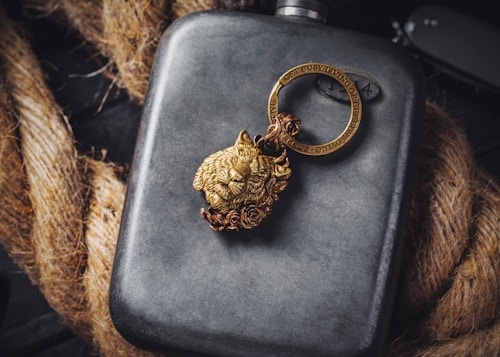 A metallic flask is placed against a backdrop of coiled rope. Resting on the flask is a detailed, gold-colored keychain featuring a lion's head and a circular ring with engraved text. The scene combines rugged textures with sophisticated details, creating a visually intriguing contrast.