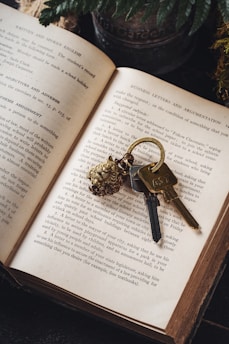 An open book lies on a dark surface with a set of ornate keys resting on one of the pages. Next to the book is a pot of ferns, partially visible. The book's text mentions topics related to writing and communication.