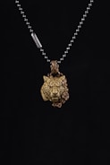 A metallic necklace featuring a pendant in the shape of a lion's head with intricate details, accompanied by a rose design beside it. The pendant is suspended from a beaded chain with a small metal tag.