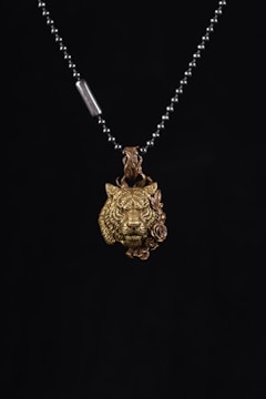 A metallic necklace featuring a pendant in the shape of a lion's head with intricate details, accompanied by a rose design beside it. The pendant is suspended from a beaded chain with a small metal tag.