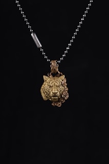 A metallic necklace featuring a pendant in the shape of a lion's head with intricate details, accompanied by a rose design beside it. The pendant is suspended from a beaded chain with a small metal tag.