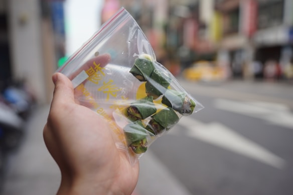 A hand holds a clear plastic bag containing several neatly wrapped green leaves in a city street setting. The background is blurred but shows a street with buildings, signs, and a taxi.