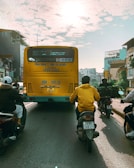 A busy urban street featuring a large yellow bus with Vietnamese writing on its rear, surrounded by several motorcyclists. The sun is shining brightly overhead through scattered clouds, casting long shadows on the road. Buildings line both sides of the street, adding an urban backdrop.