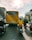 A busy urban street featuring a large yellow bus with Vietnamese writing on its rear, surrounded by several motorcyclists. The sun is shining brightly overhead through scattered clouds, casting long shadows on the road. Buildings line both sides of the street, adding an urban backdrop.