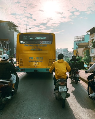 A busy urban street featuring a large yellow bus with Vietnamese writing on its rear, surrounded by several motorcyclists. The sun is shining brightly overhead through scattered clouds, casting long shadows on the road. Buildings line both sides of the street, adding an urban backdrop.