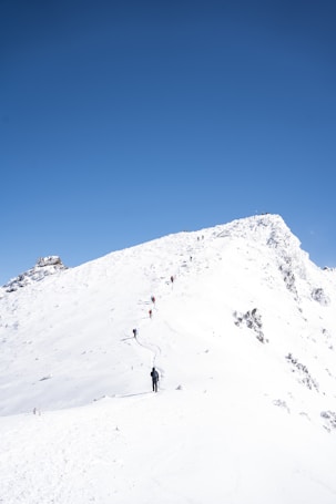 A group of people is hiking along a snow-covered mountain ridge under a clear blue sky. The slope is steep and the path is narrow, with footprints leading up the hill. The terrain is rugged, with patches of rocks peeking through the snow.