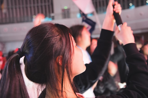 A young person with dark hair tied in a ponytail is seen in profile, holding an item that resembles a phone or a camera. The setting appears to be indoors, with other people blurred in the background, suggesting a crowded or lively environment.